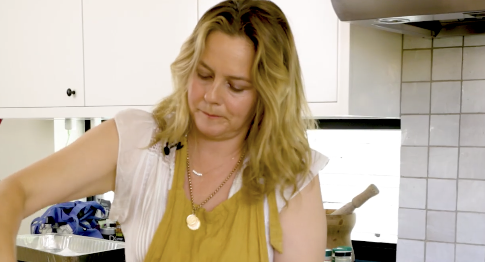A woman with shoulder-length blonde hair wears a yellow apron while preparing food in a kitchen.