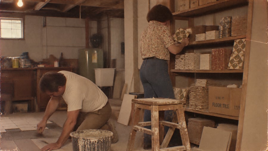 A man kneeling on a basement floor lays tile while a woman stands at a wooden shelf selecting patterned tiles.