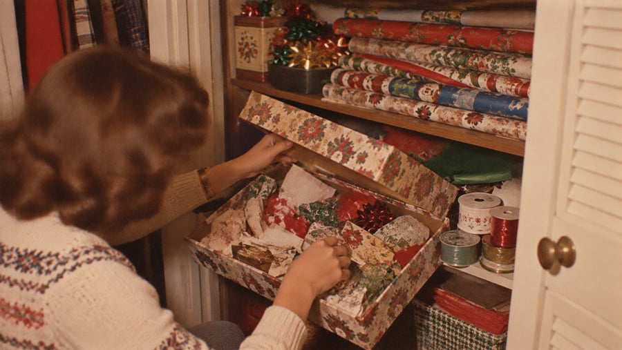 A person reaching into a closet to open a floral gift box filled with wrapping supplies, with rolls of gift wrap and ribbon stored on shelves above.