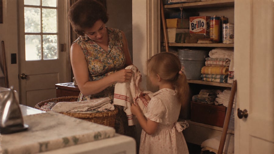 A woman in a floral dress and a young girl holding a white cloth with a red stain stand in a pantry.