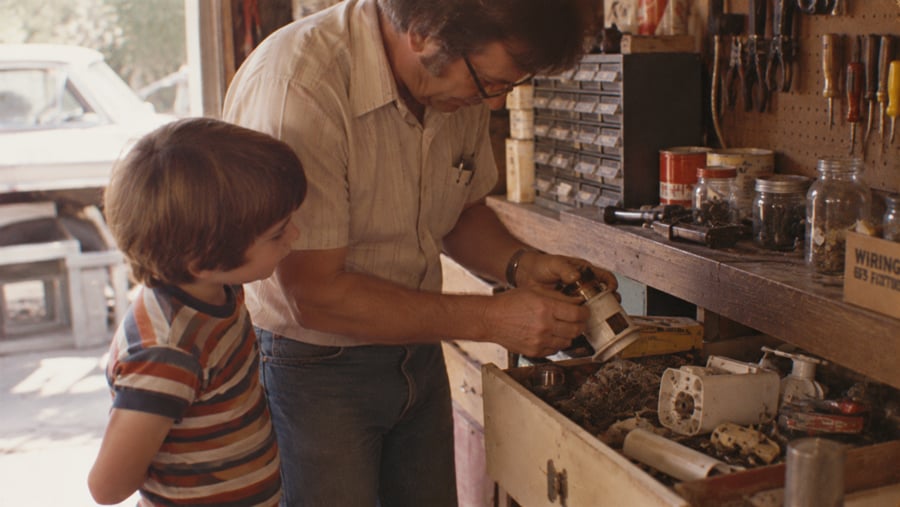 An adult man and a young child look at a small mechanical part together in a workshop.