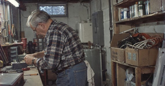 An older man in a plaid shirt and glasses works on electrical components at a workbench in a workshop.