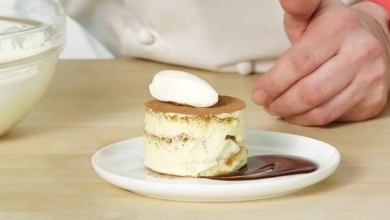 A small, round tiramisu cake topped with a dollop of cream sits on a plate next to a swirl of chocolate sauce.
