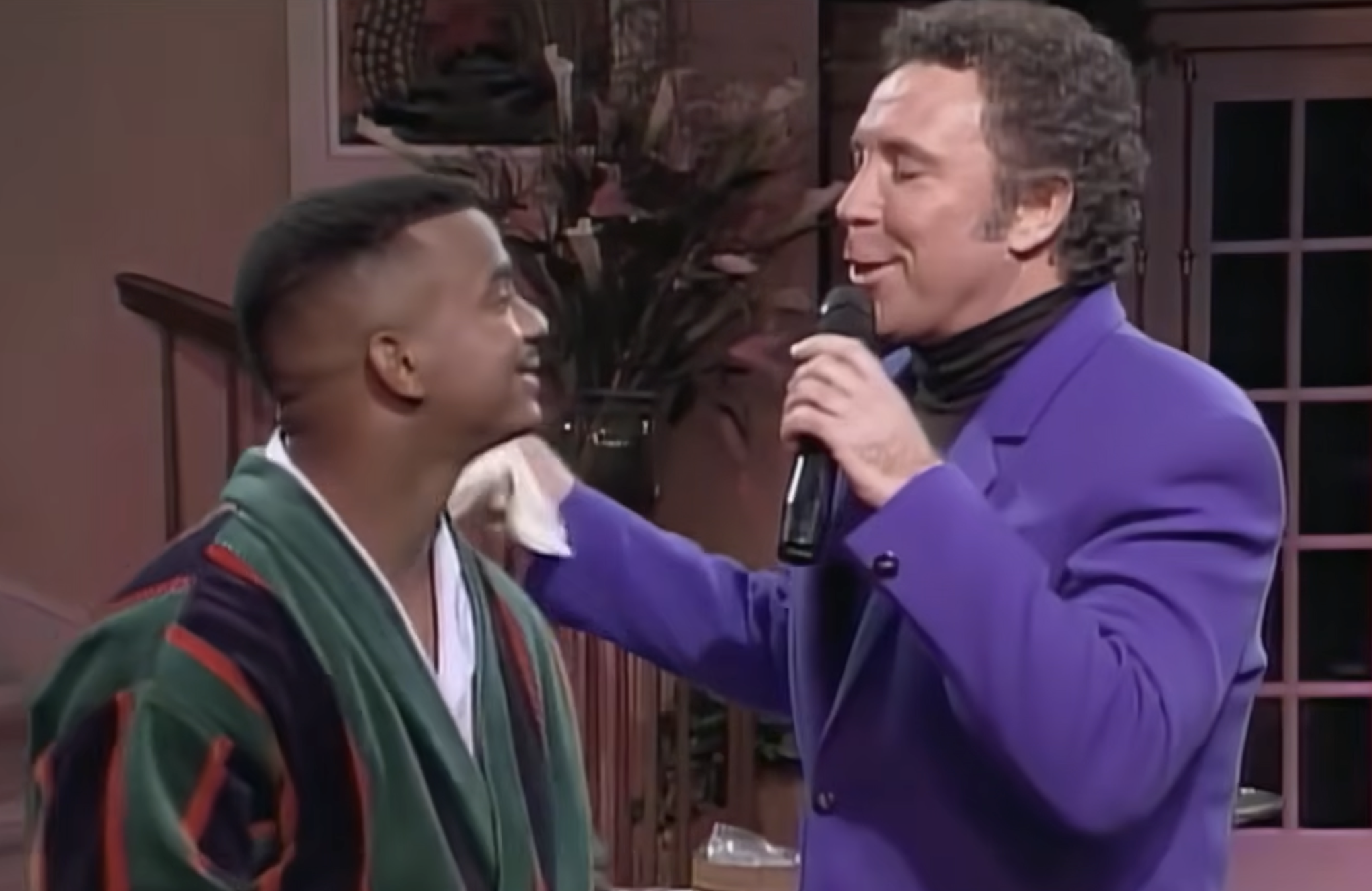 Tom Jones in a purple blazer sings into a microphone while wiping the cheek of Alfonso Ribeiro, who is wearing a robe.