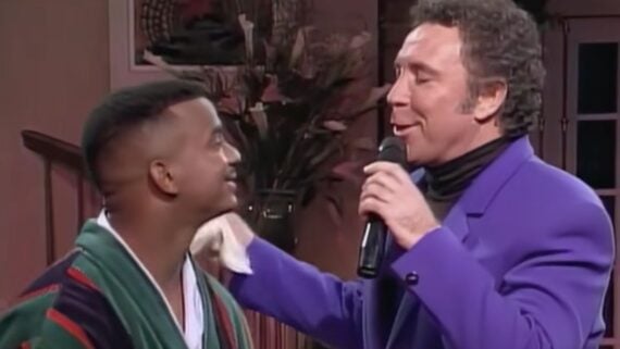Alfonso Ribeiro, wearing a robe, smiles at Tom Jones, who is singing into a microphone while wearing a purple blazer.