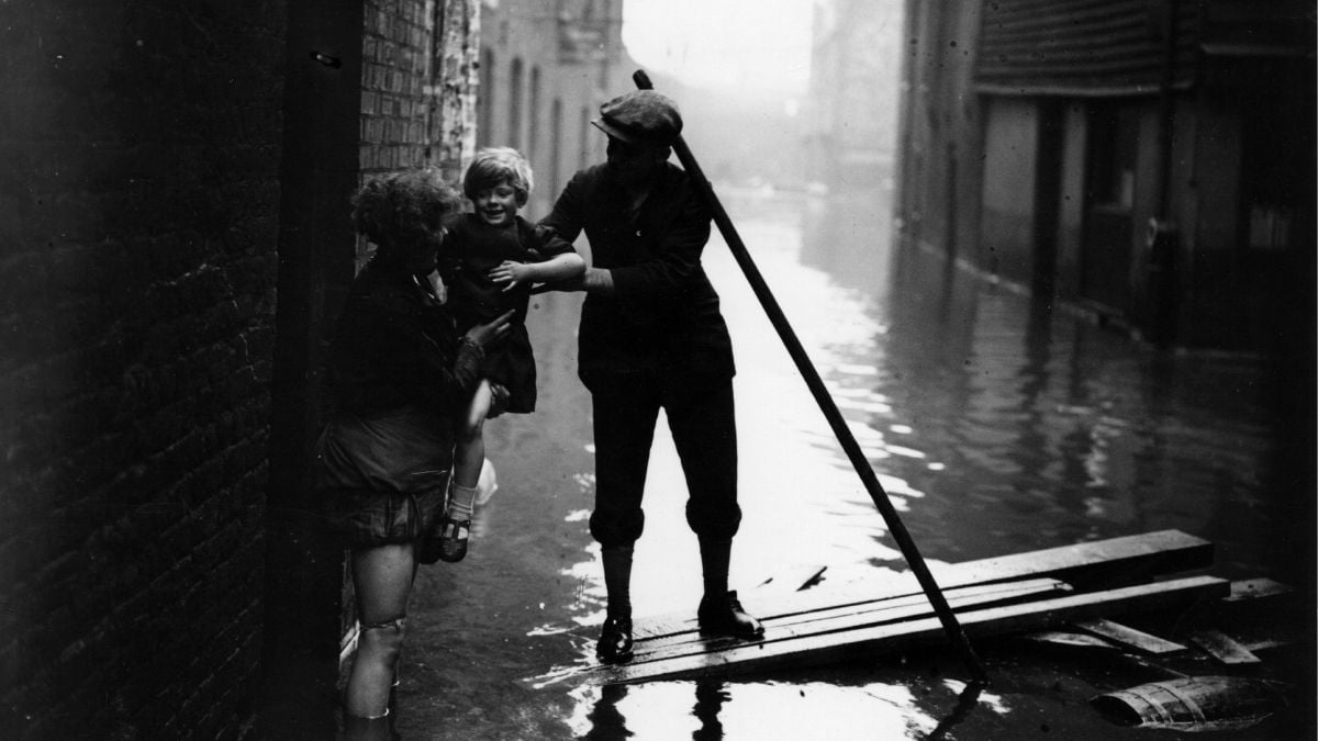 A person stands in a flooded street, helping a child to safety while another adult sits nearby, all surrounded by water and debris.