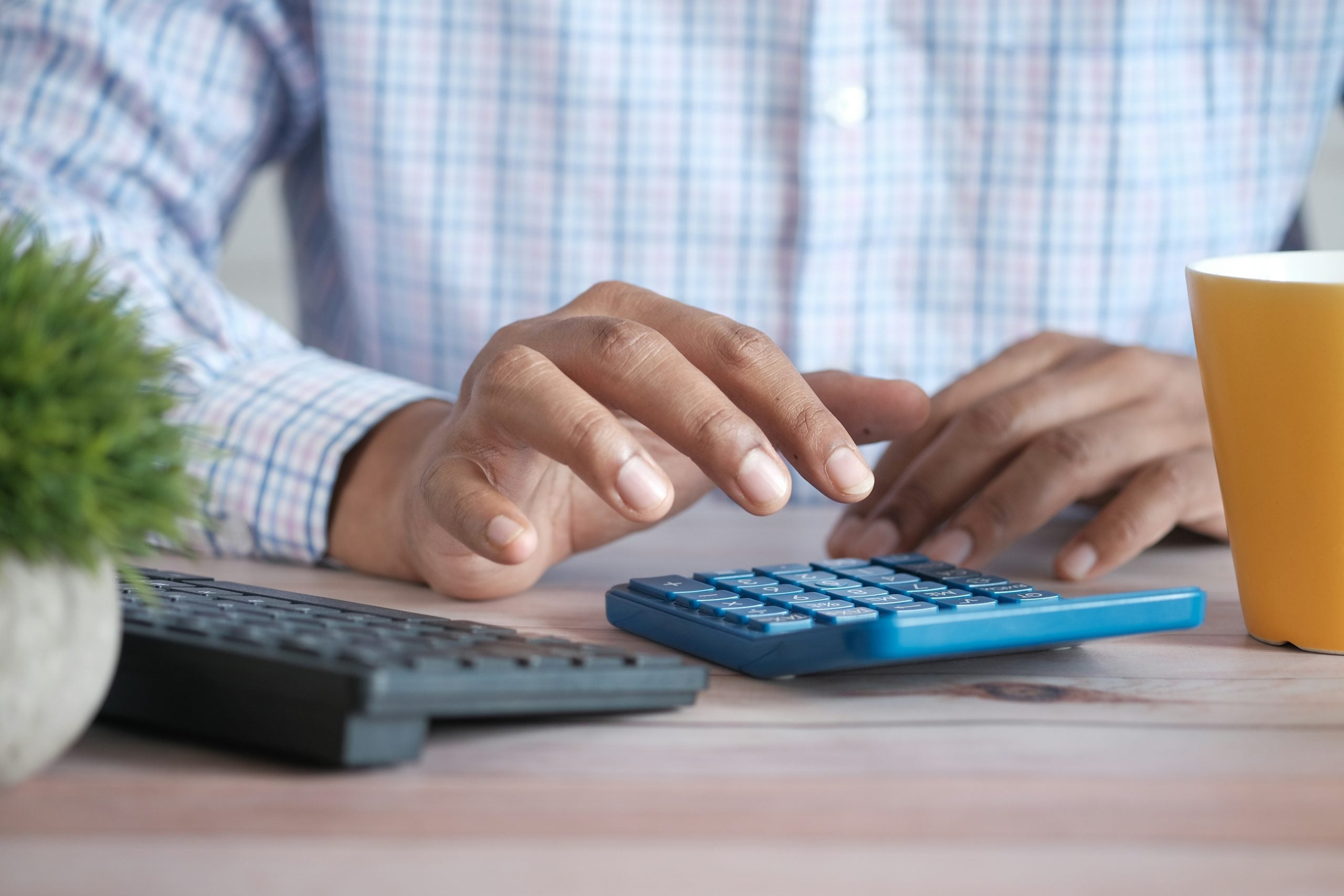 A person in a checkered shirt uses a blue calculator on a desk next to a keyboard and a yellow mug.