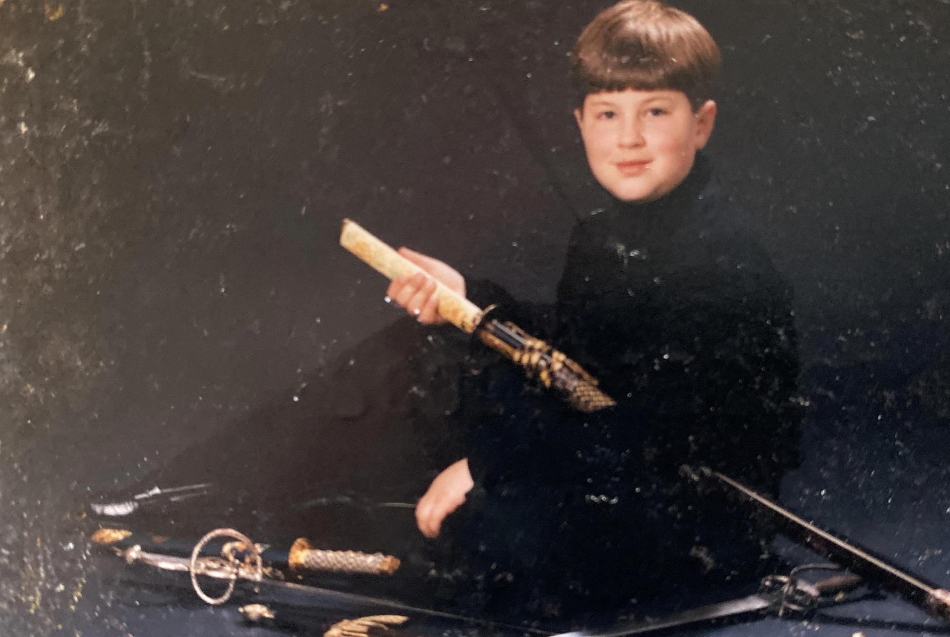 A young boy in a black turtleneck holds a decorative dagger, with a sword resting on the floor in front of him.