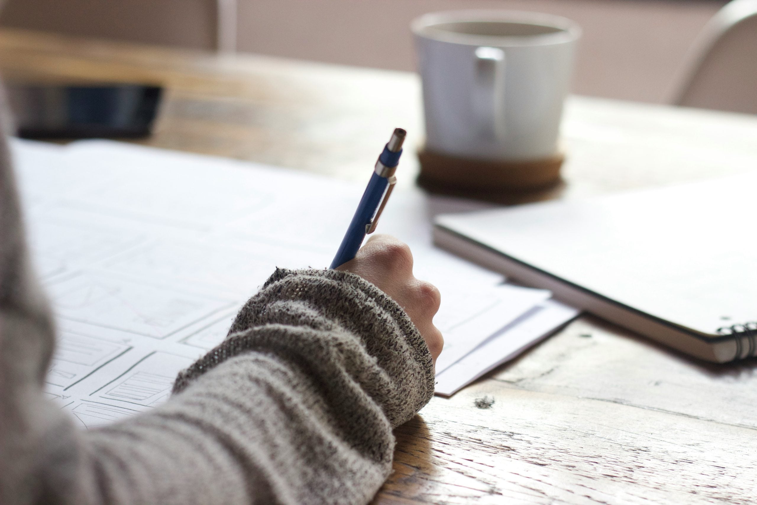 A person holding a pen over a sheet of paper with sketches on a wooden table next to a coffee mug.