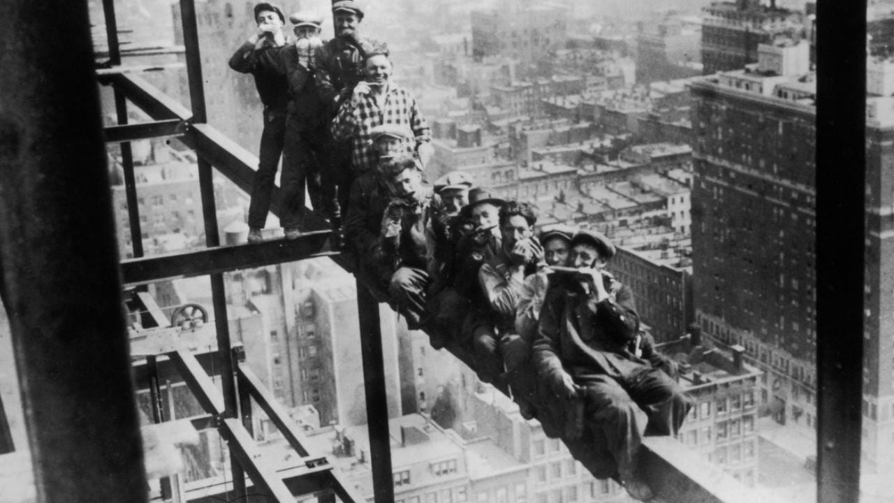 Eleven construction workers take a break, sitting and standing on a steel beam high above a city skyline, with buildings and streets visible far below.