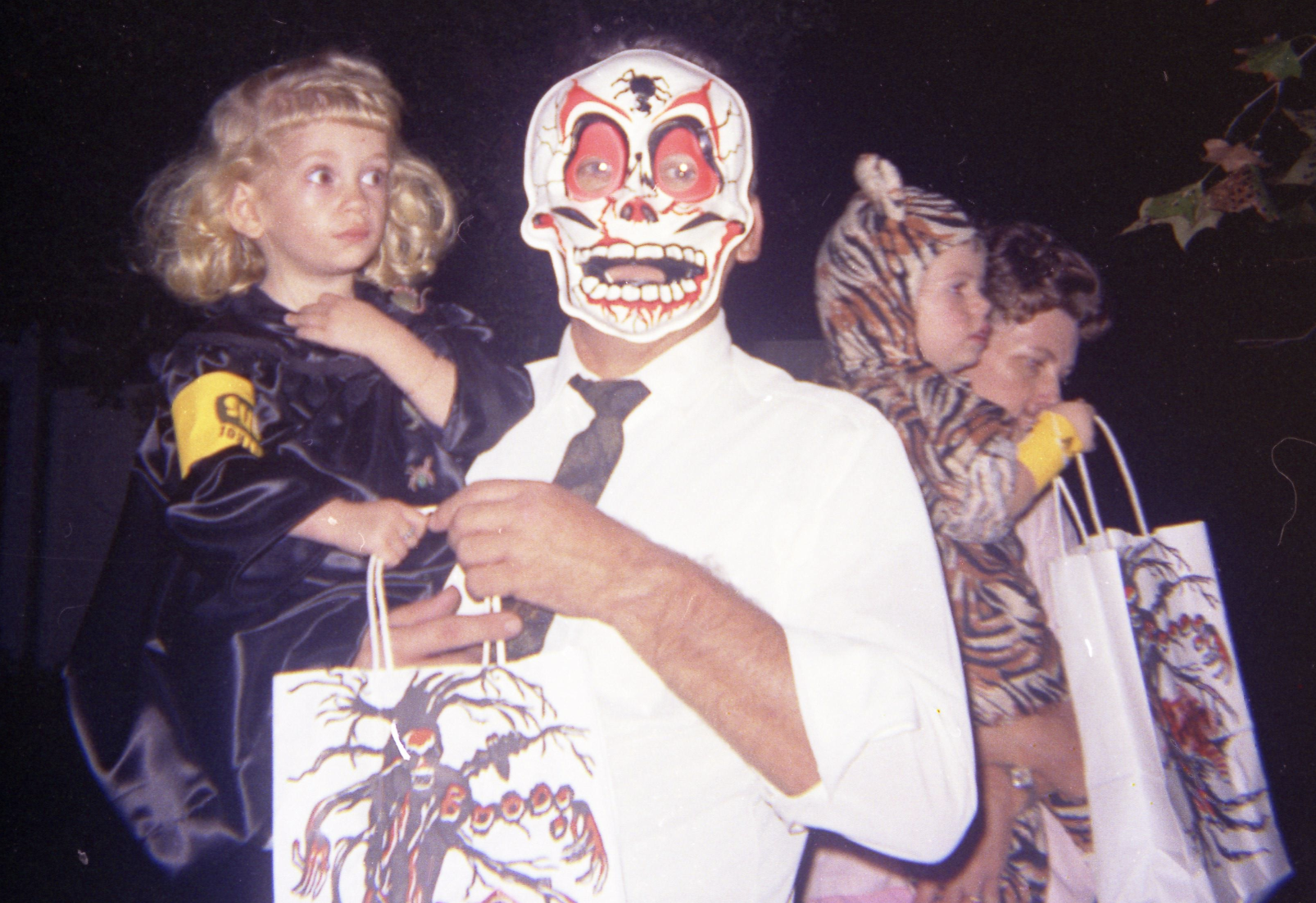 An adult wearing a skull mask holds a young girl in a costume, while a woman beside them carries a child in a tiger costume, all holding trick-or-treat bags.