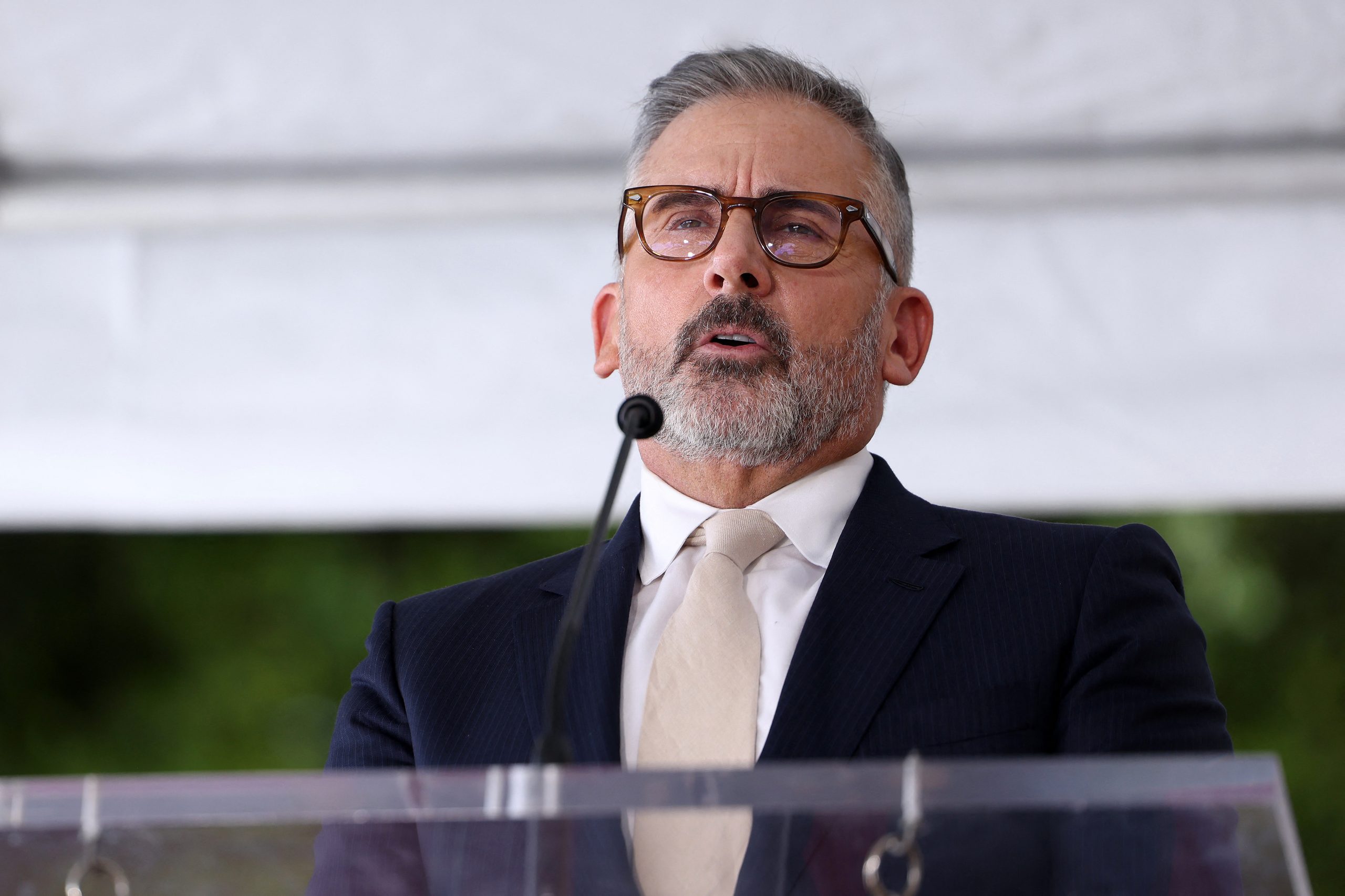 Steve Carell wearing a suit, tie, and glasses speaks at a podium.