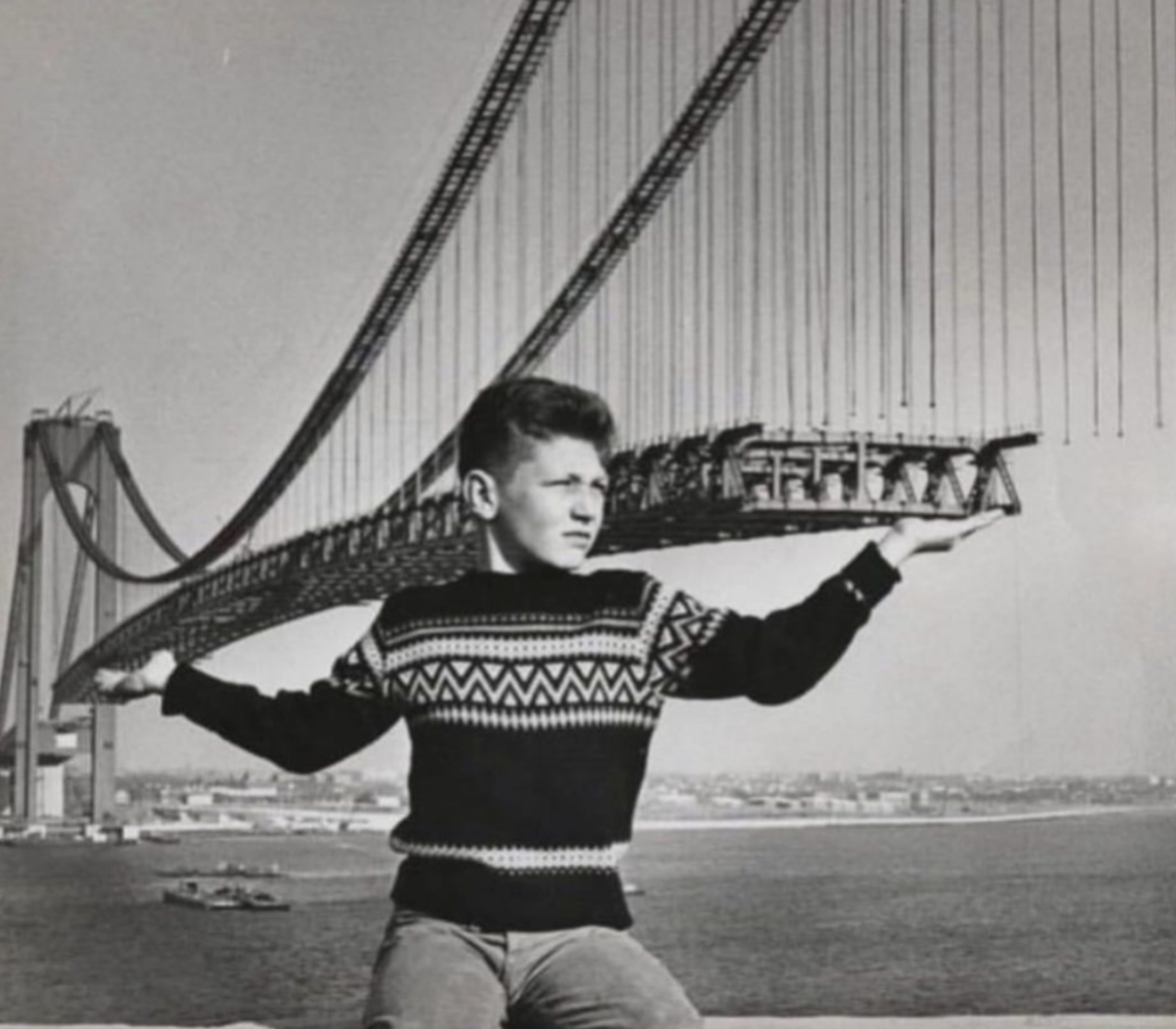 A boy poses in the foreground as if holding up a section of the Verrazzano-Narrows Bridge under construction.