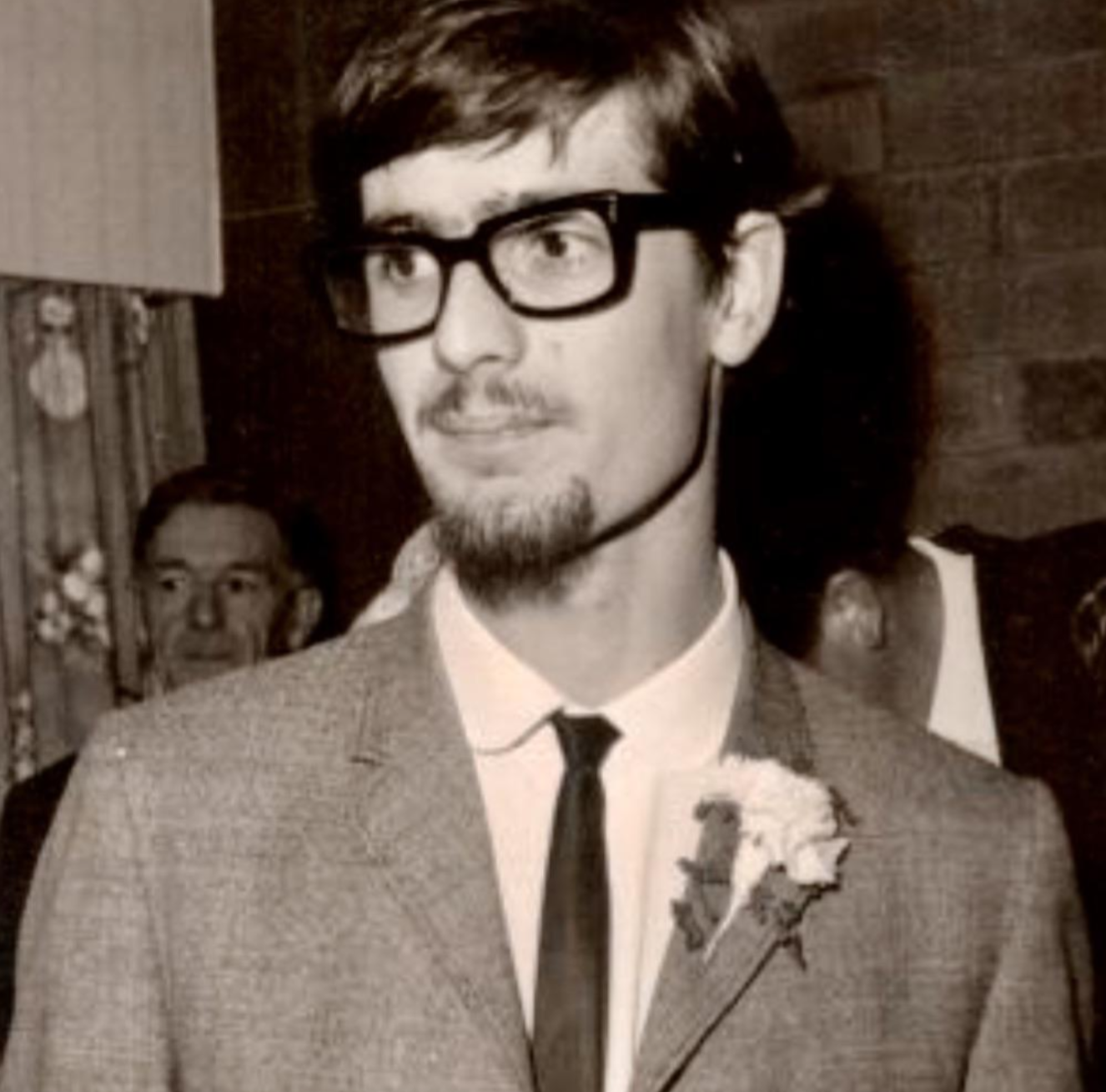 A young man with dark hair, a goatee, and thick-rimmed glasses wearing a suit, white shirt, and tie with a flower boutonniere.