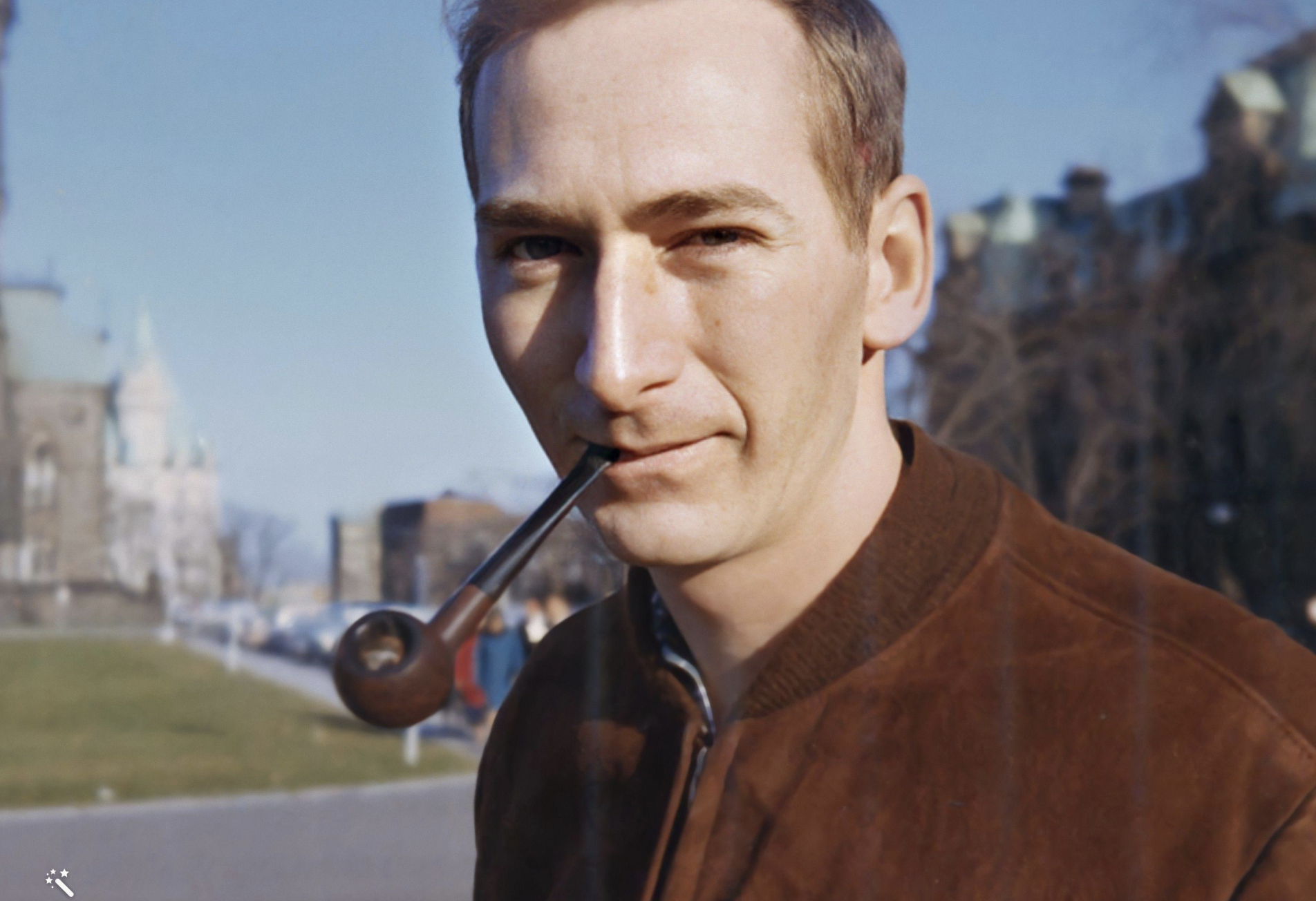 A man in a brown jacket looking at the camera while holding a pipe in his mouth.