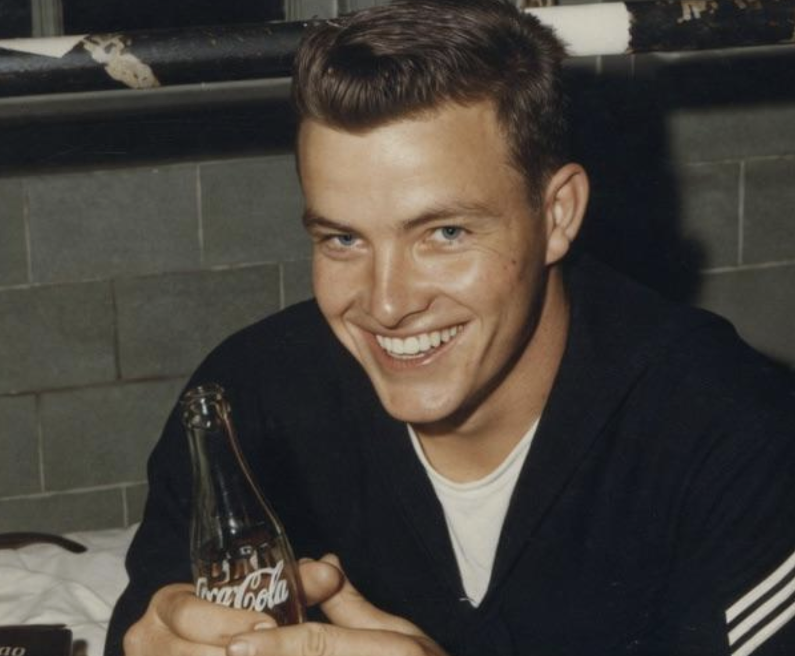 A smiling man in a navy uniform holds a glass bottle of Coca-Cola.