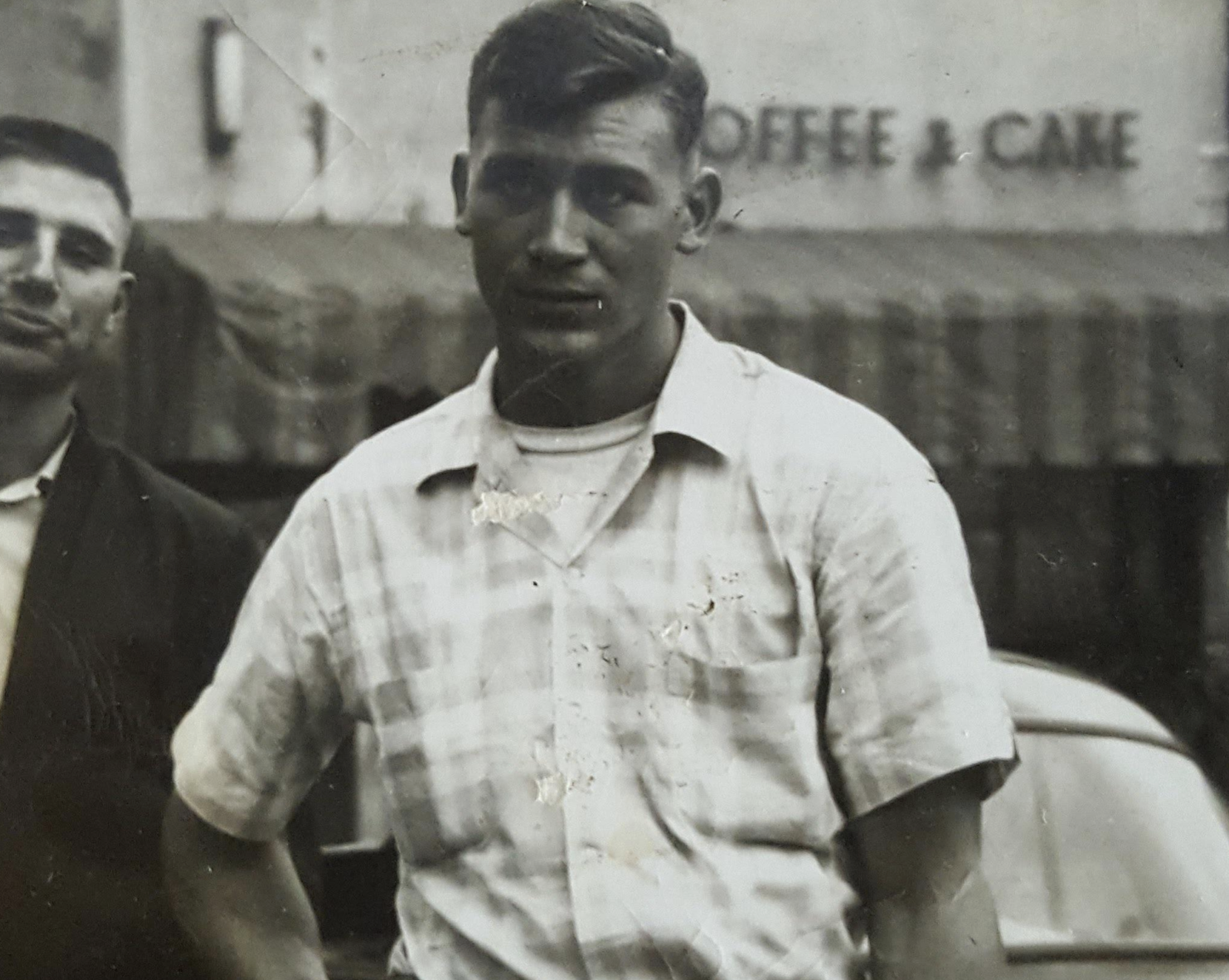 A black and white vintage photograph of two young men standing in front of a building with the words “Coffee & Cake” visible on the storefront.