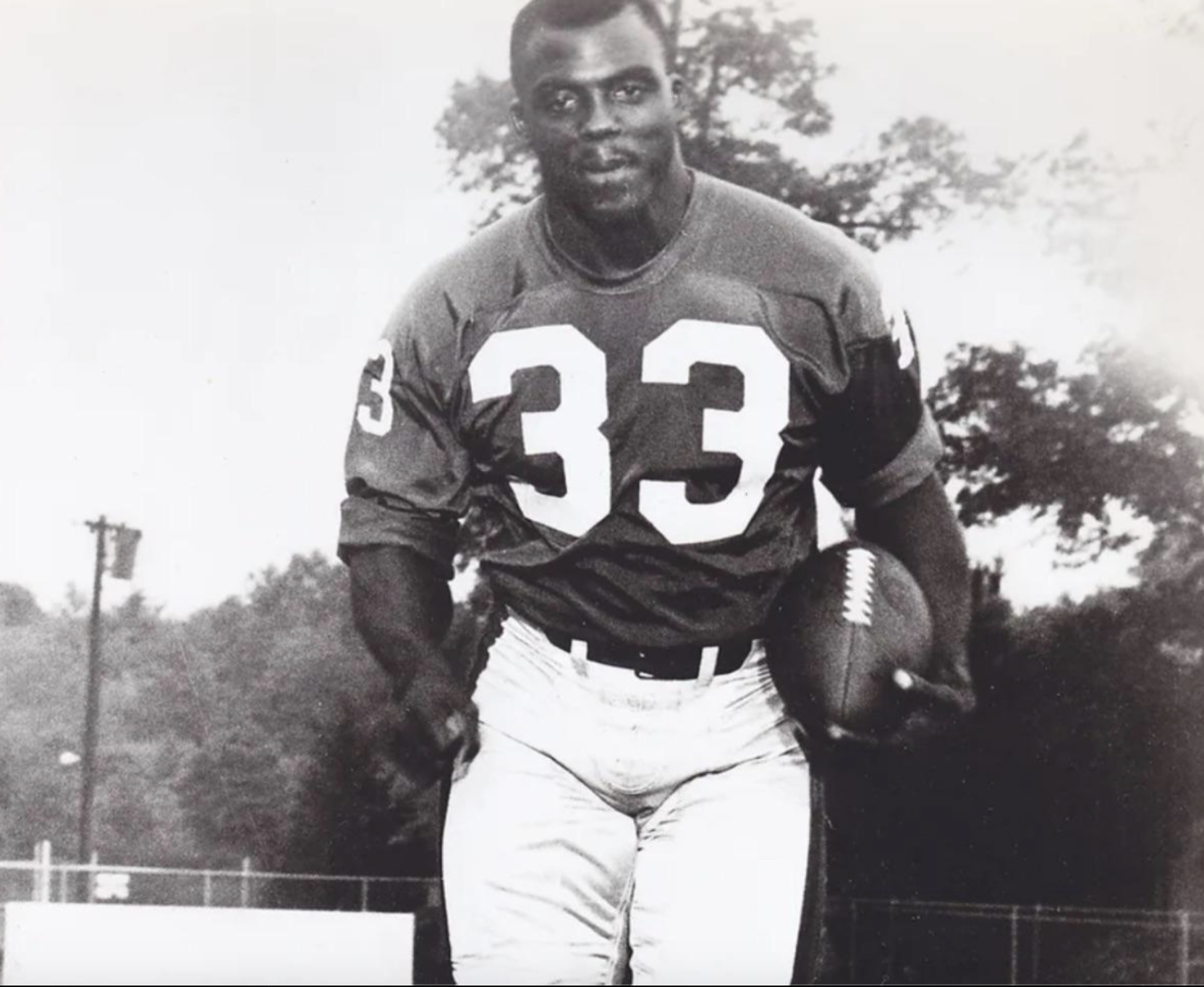 A black-and-white photo of a football player wearing a jersey with the number 33, carrying a football under his arm.