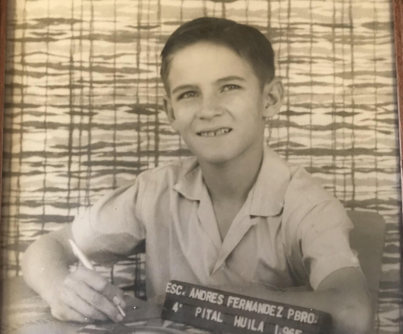 A young boy in a collared shirt sits at a desk with a nameplate that reads “ESC. ANDRES FERNANDEZ PBRO. 4° PITAL HUILA 1965.”