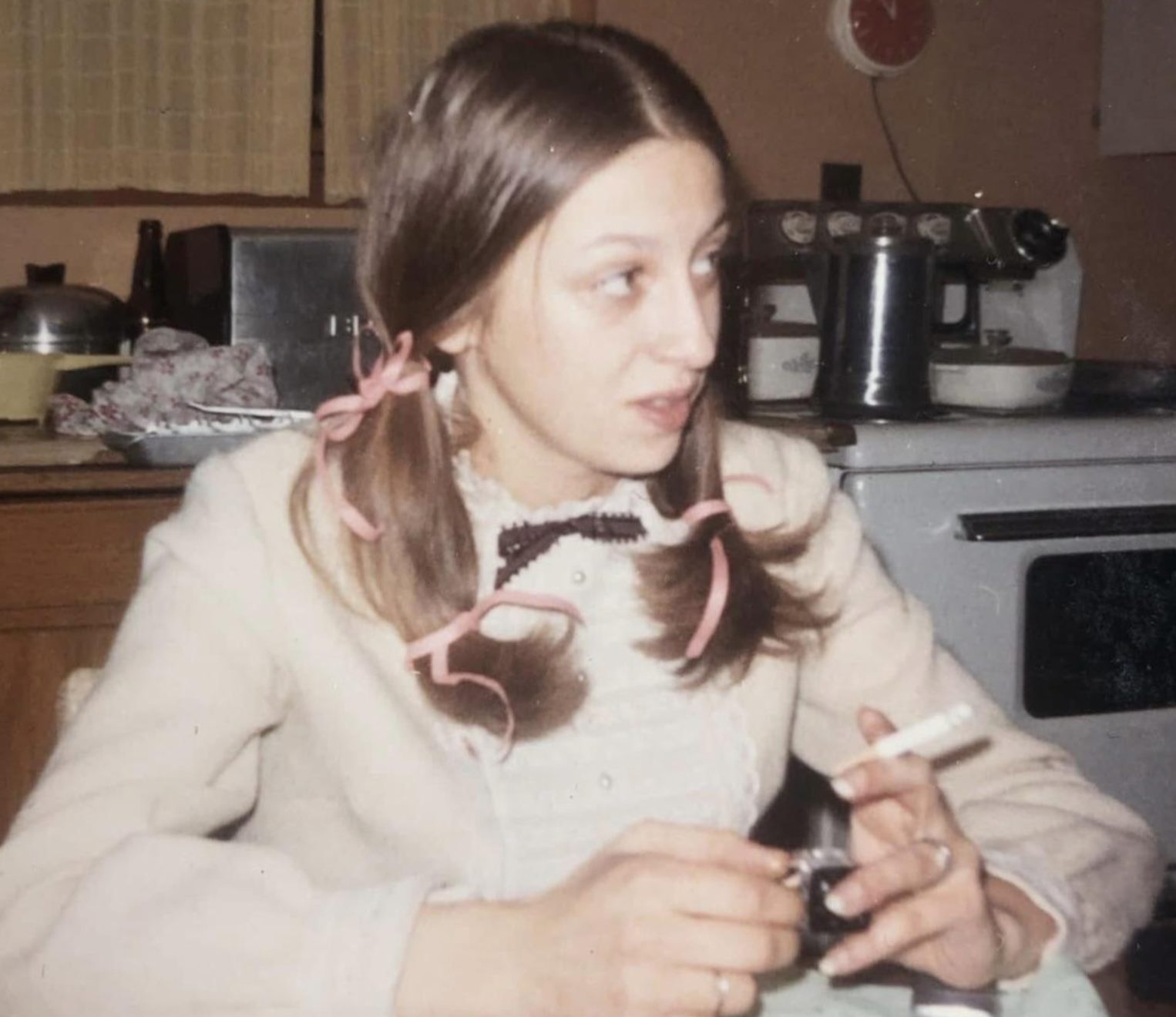 A young woman with pigtails tied with pink ribbons sits at a table holding a lit cigarette.