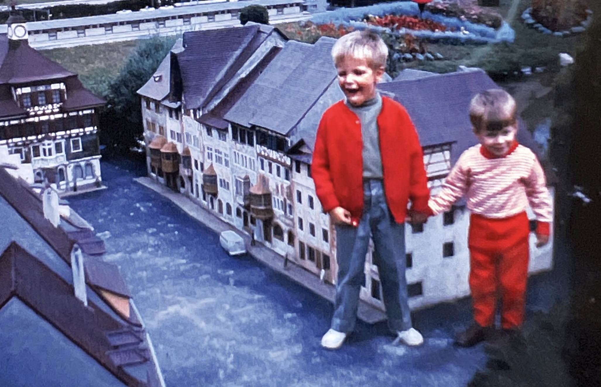 Two children holding hands while standing in front of a miniature model of a riverside town.