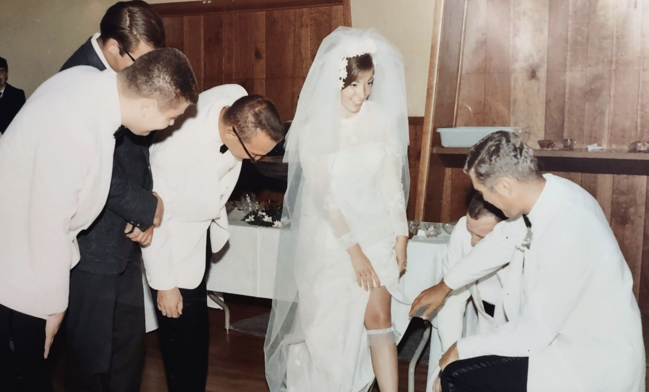 A bride in a white wedding gown and veil lifts her skirt to show a garter on her leg to a group of men in suits.