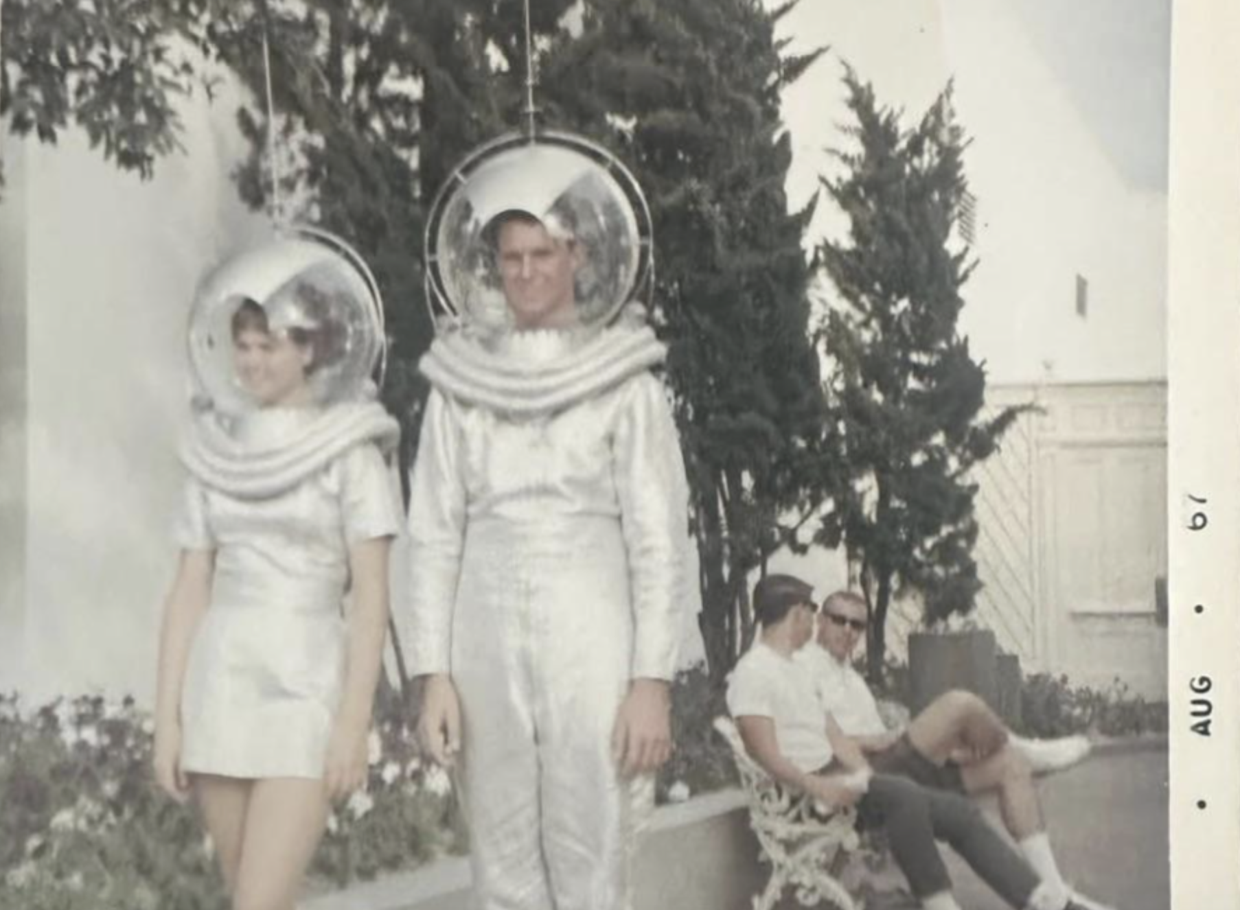 A woman and man in silver astronaut costumes with spherical helmets stand outside, while two men sit on a bench nearby.