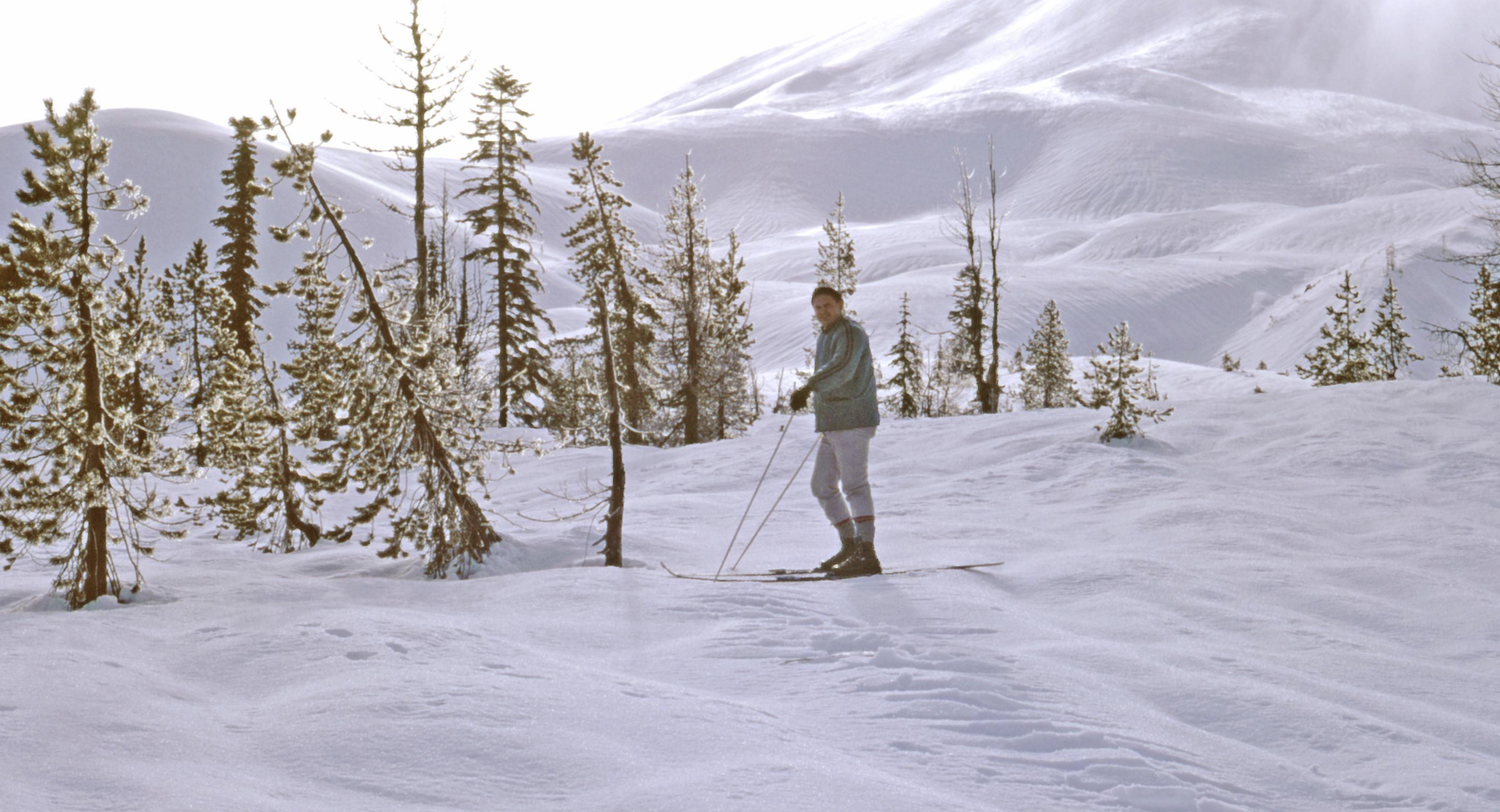 A person in a light jacket and pants cross-country skis through a snowy, wooded landscape toward a large, snow-covered hill.
