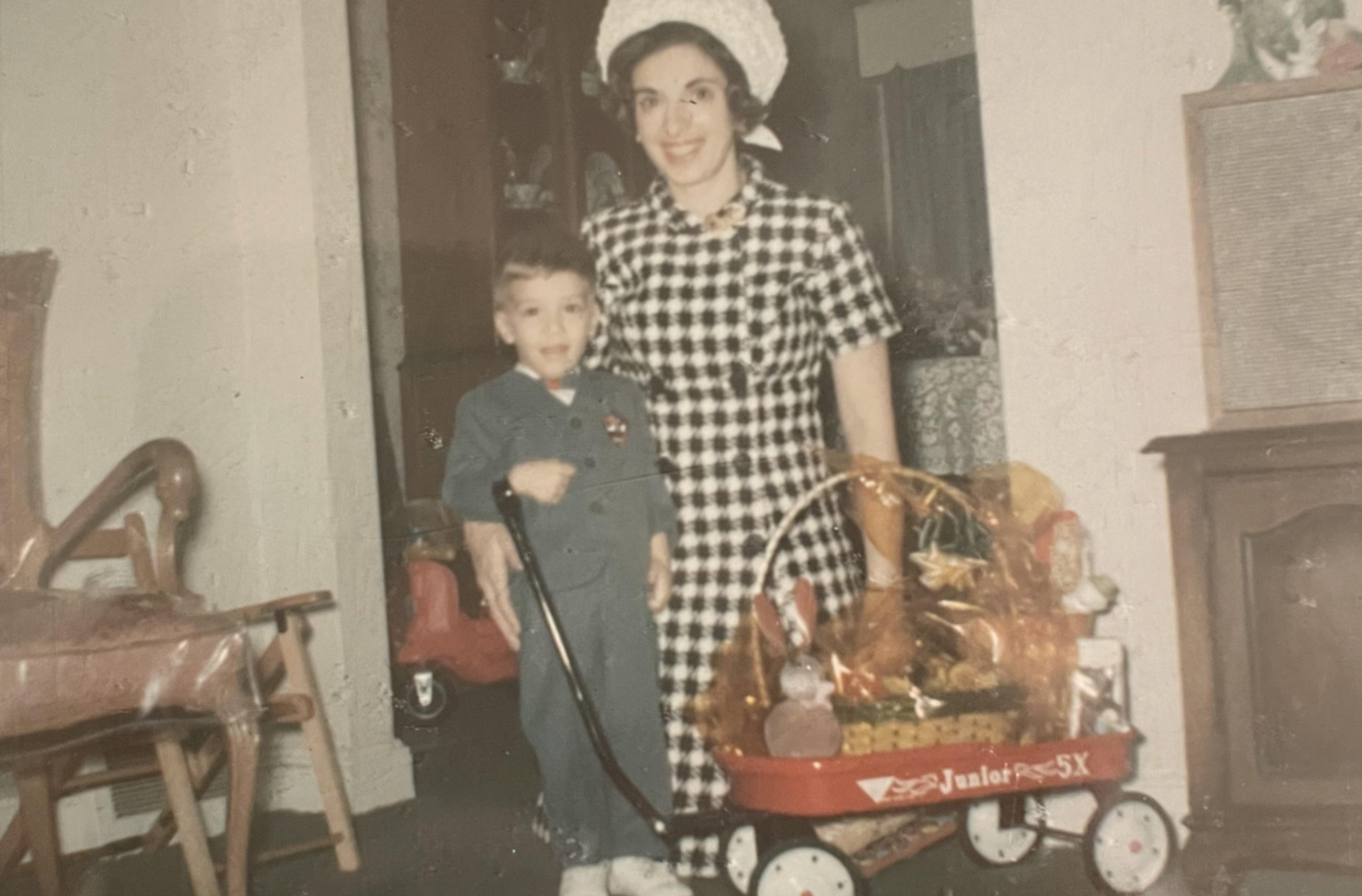 A woman in a gingham dress and a young boy in a suit stand together next to a red toy wagon filled with gifts.