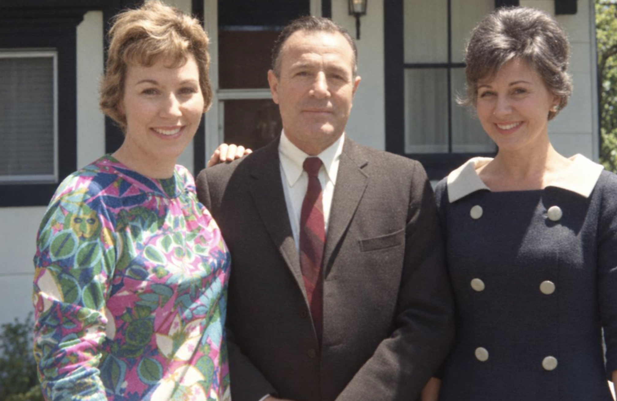 A man in a suit and tie standing between two smiling women outdoors.