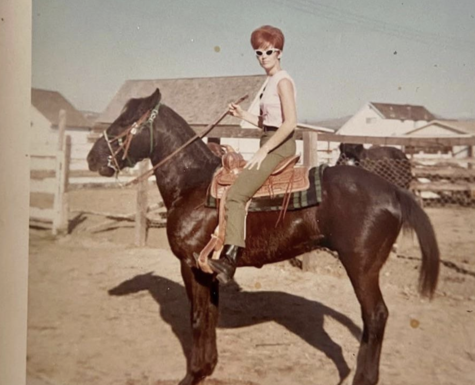 A woman with a red beehive hairstyle and sunglasses sits on a saddled horse in a corral.