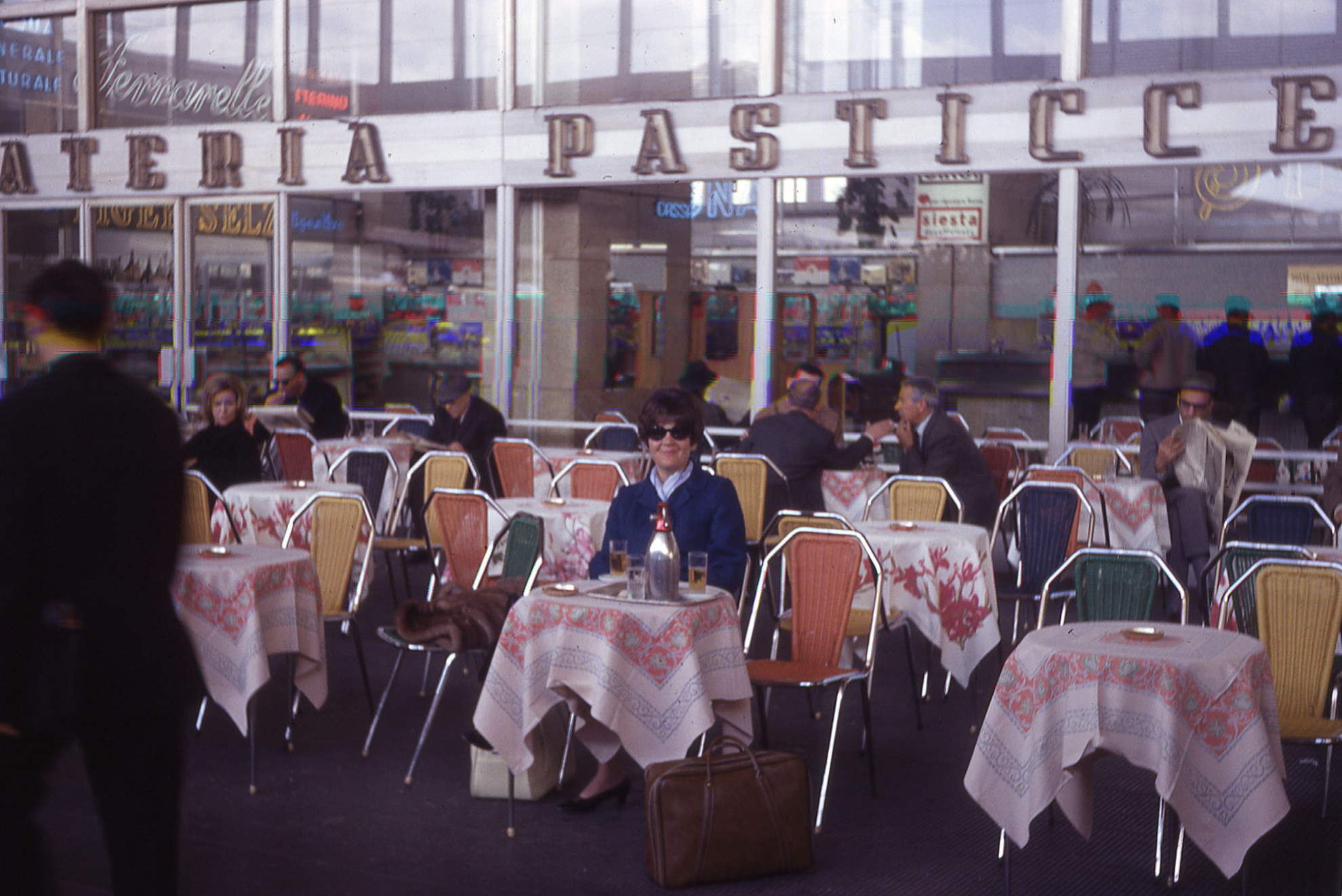 A woman in sunglasses sits at a table on the outdoor patio of a pastry shop, with other patrons dining in the background.