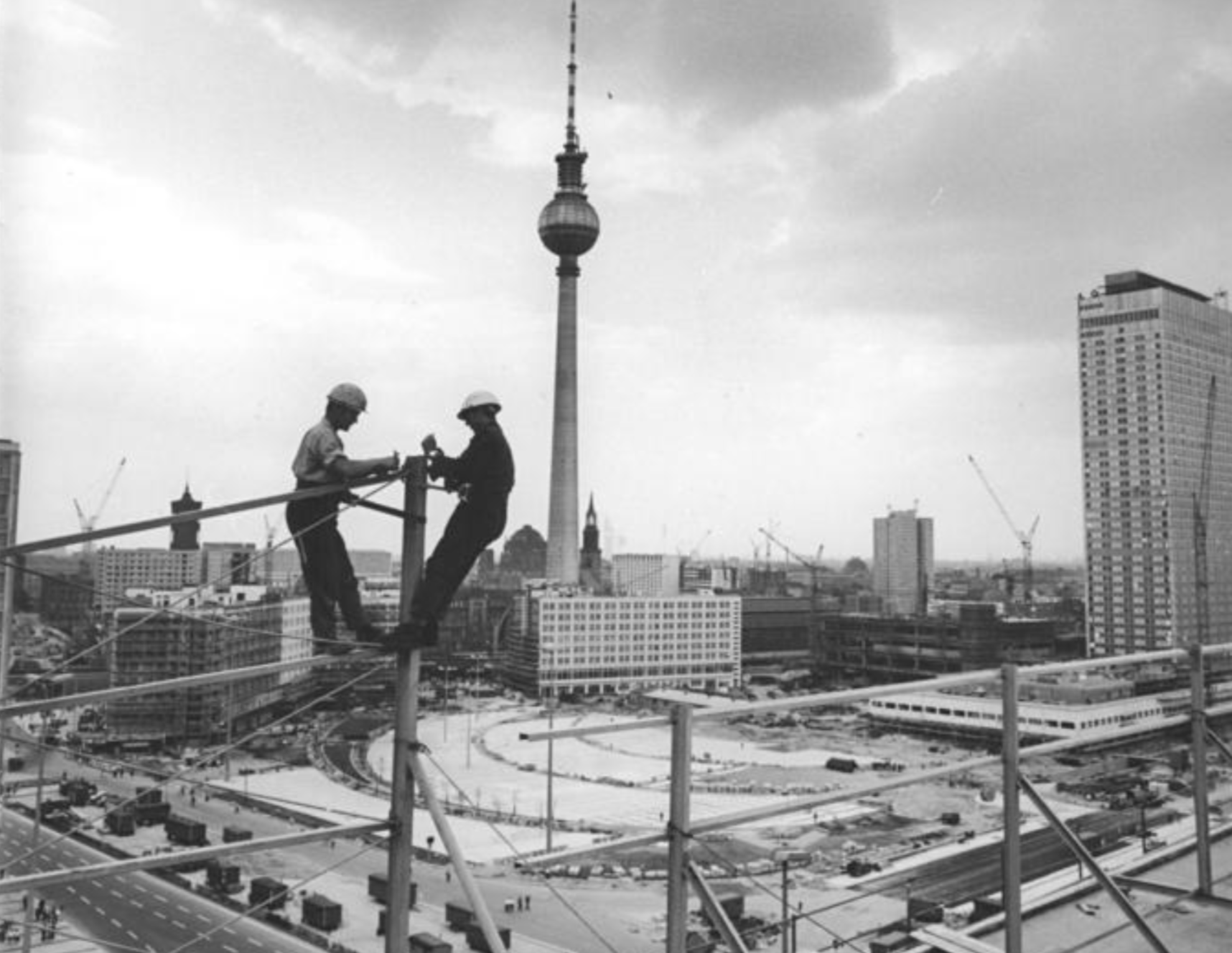 Two construction workers stand on scaffolding high above Berlin, with the TV Tower visible in the background.