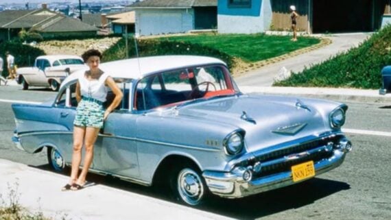 A woman in a white top and patterned shorts leans against a silver 1957 Chevrolet Bel Air parked on a suburban street.