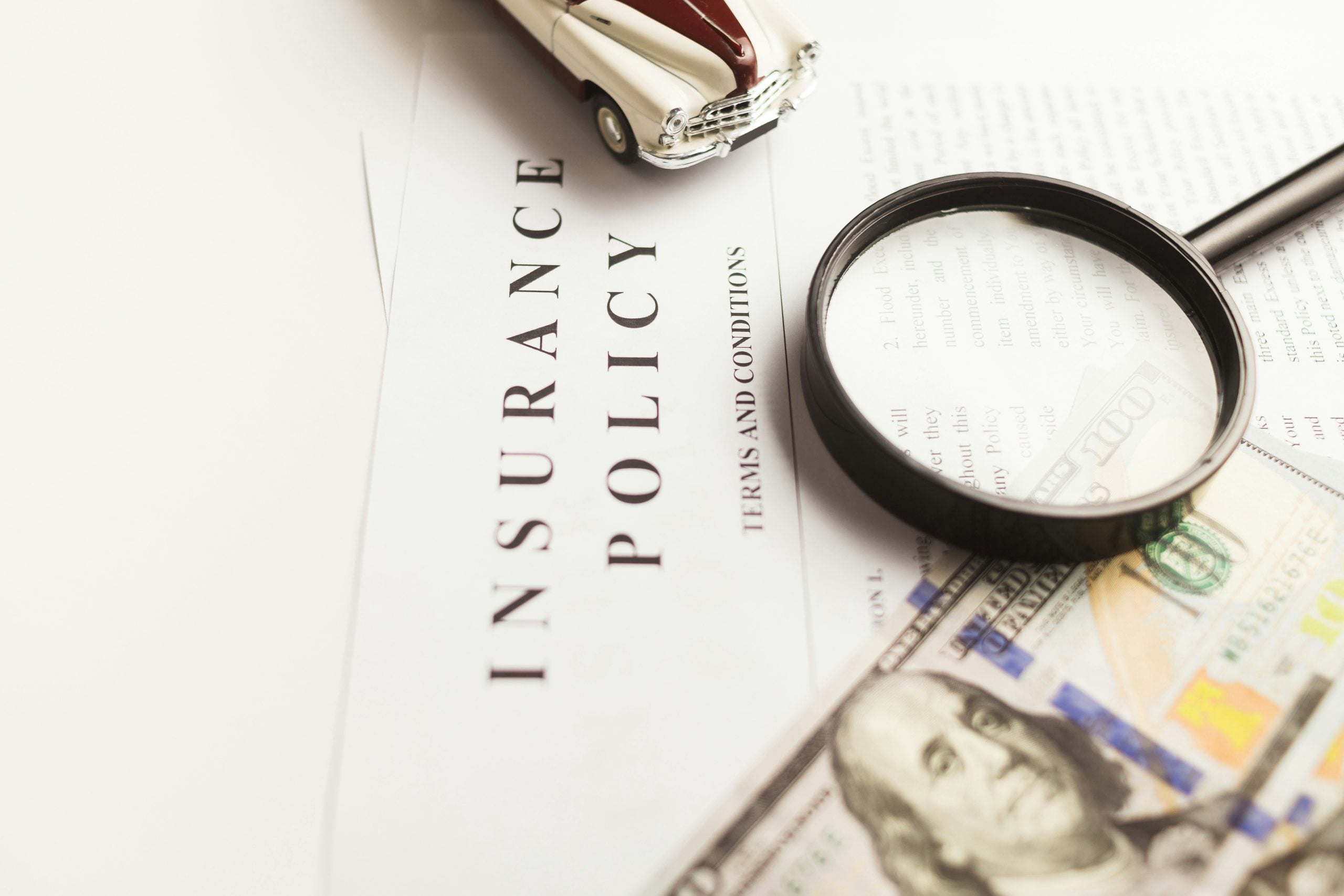 An insurance policy document with a magnifying glass, a toy car, and a hundred-dollar bill.
