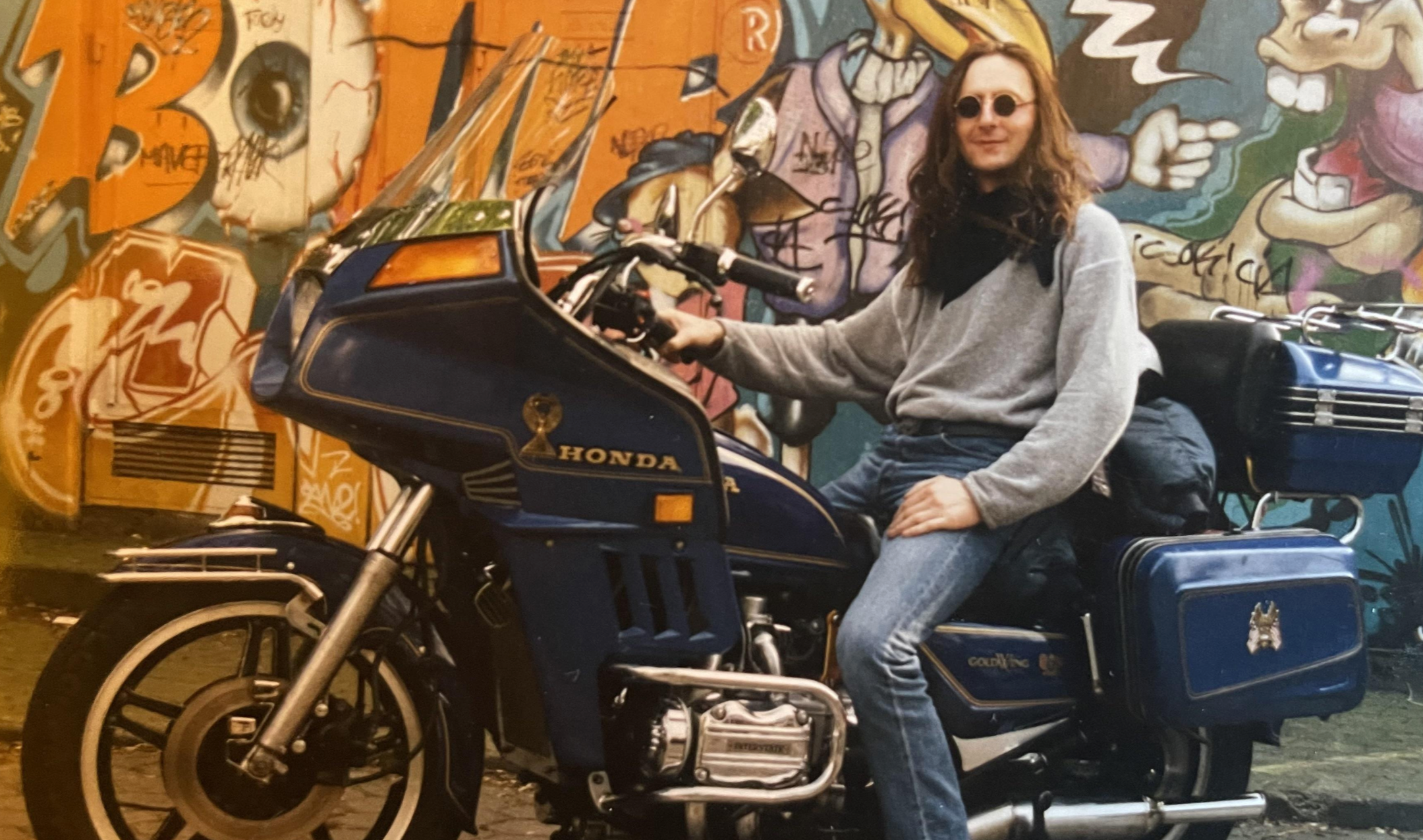 A person with long hair and round sunglasses sitting on a blue Honda Gold Wing motorcycle in front of a graffiti-covered wall.