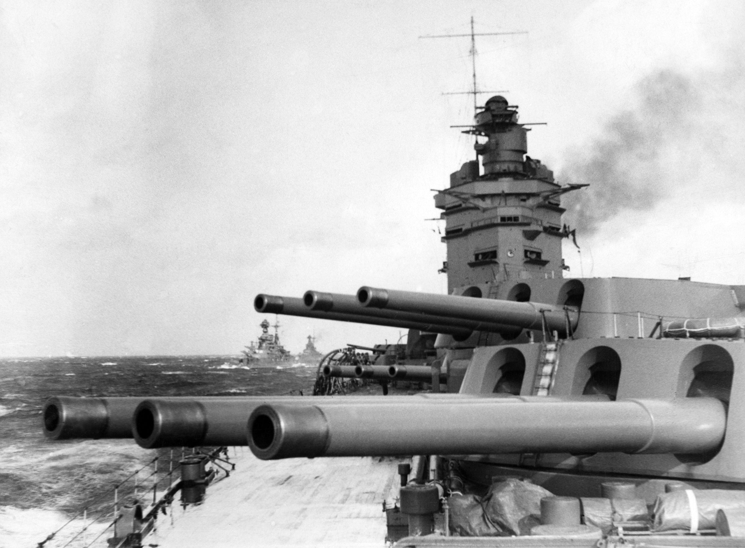View from the deck of a battleship showing large naval guns in the foreground, with another battleship visible in the distance across a choppy sea under a cloudy sky.