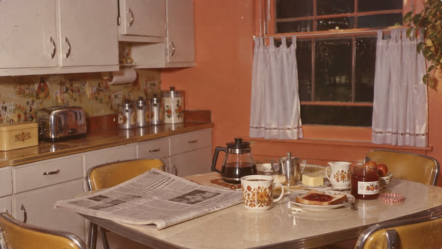 A vintage kitchen with peach walls, floral curtains, and a table set with coffee, toast, jam, fruit, and a newspaper. White cabinets, canisters, and a toaster are visible in the background.