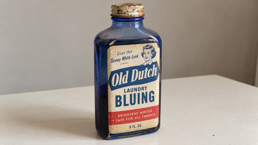 A vintage blue glass bottle labeled “Old Dutch Laundry Bluing” sits on a white surface. The label features a woman’s face and advertises that it “brightens whites” and is “safe for all fabrics.”