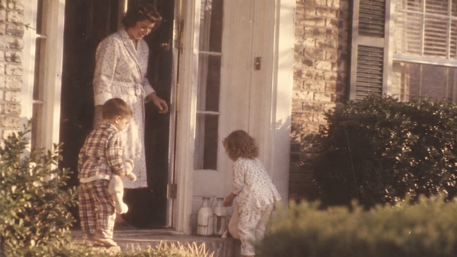 A woman in a robe stands at a door while two young children in pajamas pick up glass milk bottles from the porch steps outside a brick house.