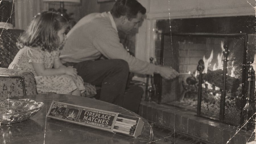 A man and a young girl sit by a lit fireplace. The man tends to the fire while the girl watches. A box of fireplace matches and an ashtray are on a nearby table. The photo has a vintage, black-and-white look.