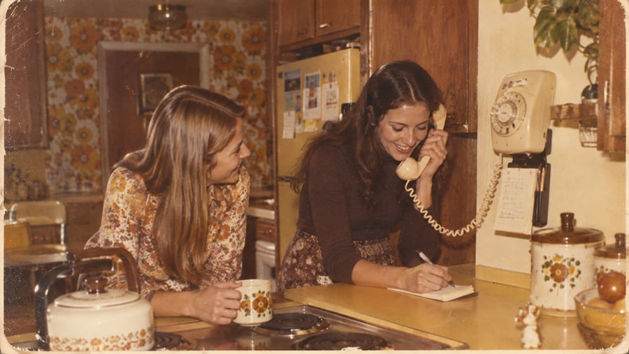 Two women in a vintage kitchen, one talking on a wall-mounted rotary phone and writing a note, the other holding a mug. The kitchen has brown and yellow floral decor and an old-style refrigerator.
