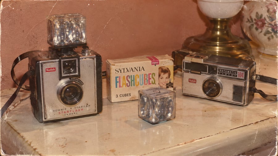 Two vintage Kodak cameras, a Sylvania Flashcubes box, and several flashcubes are displayed on a table beside a lamp and a floral vase.