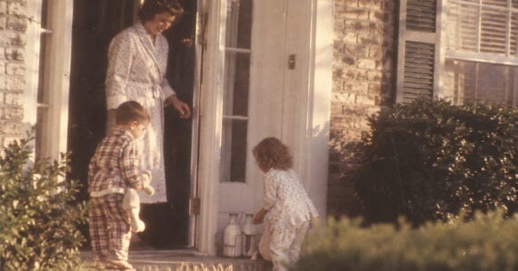 A woman in a robe stands at a door with two small children in pajamas. The children look at several glass milk bottles placed on the doorstep near the bushes.
