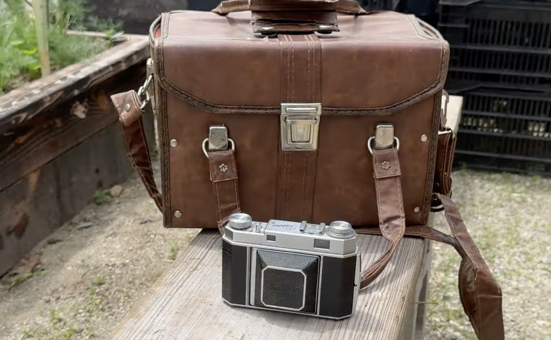 A vintage 35mm camera sits on a wooden surface in front of a brown leather camera bag.