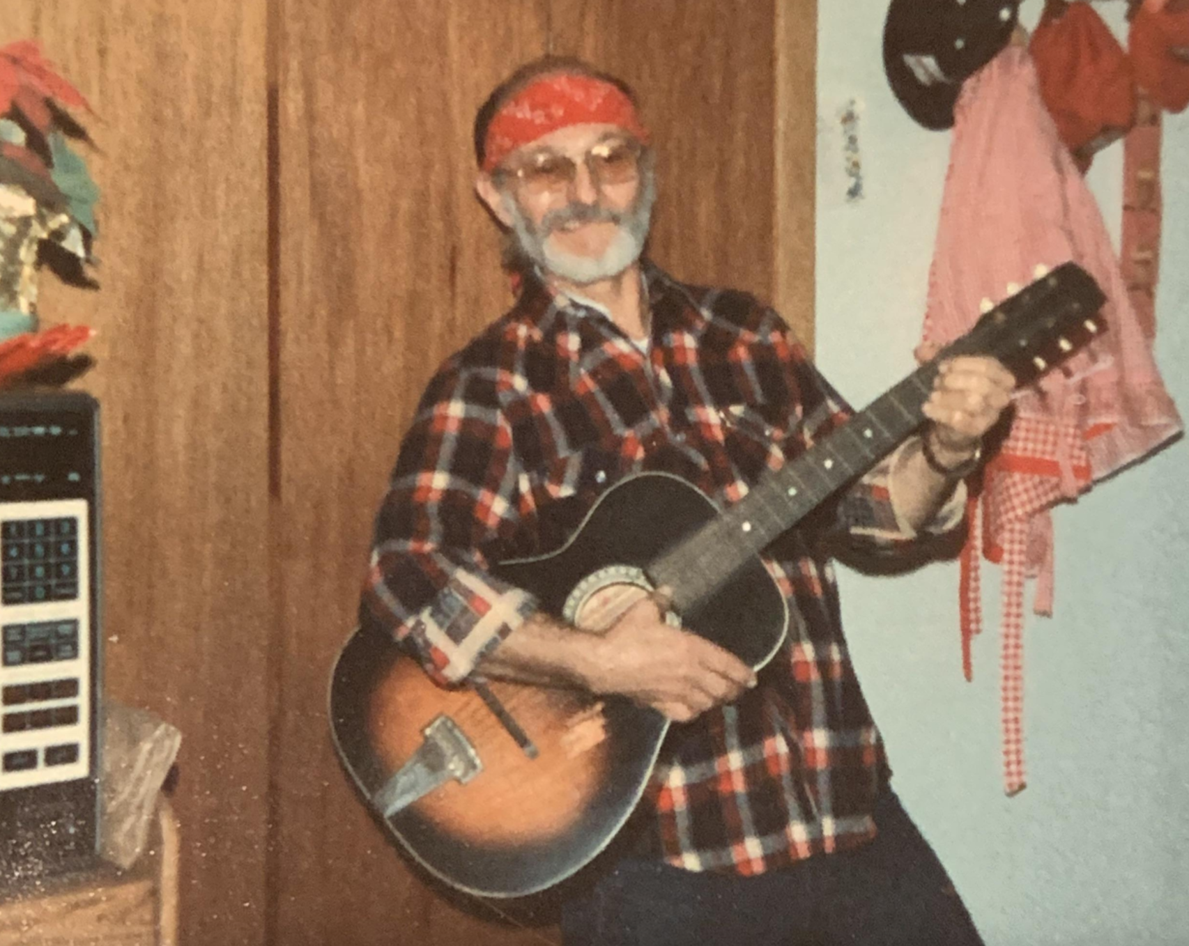An older man with a white beard and a red bandana holds and plays an acoustic guitar.