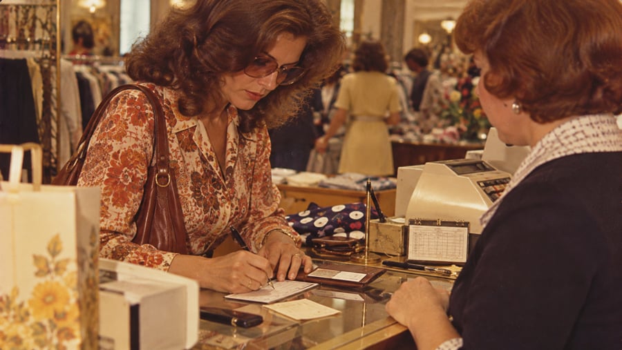 A woman wearing sunglasses writes a check at a store counter while a cashier stands nearby. Shopping bags and a cash register are visible on the counter.