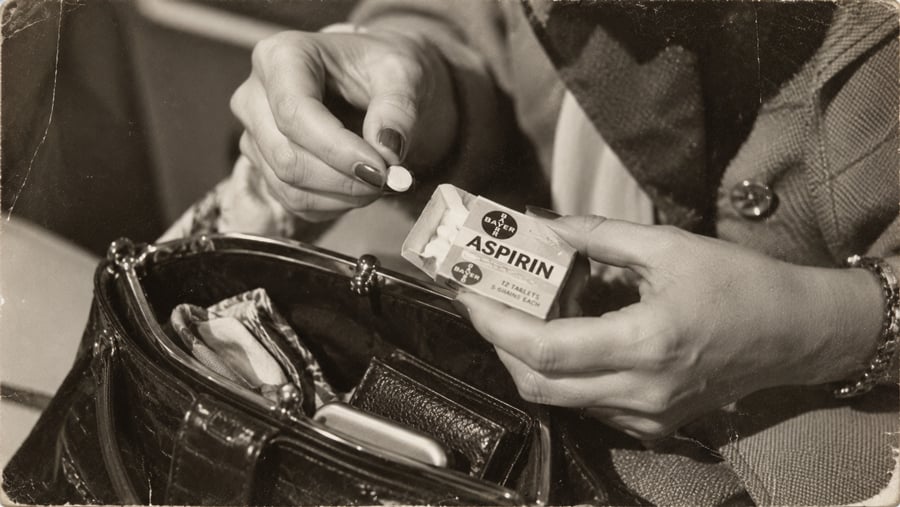 A person holds a round aspirin tablet in one hand and an open box labeled “Aspirin” in the other, above an open handbag containing a wallet and other items. The image is in black and white.