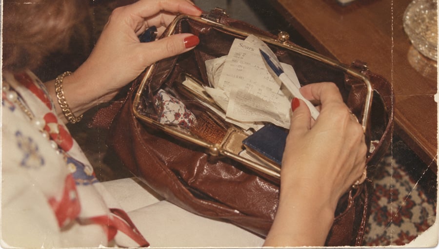 A person with red nail polish holds an open brown purse filled with receipts, papers, a pen, and other small items, sitting at a table.