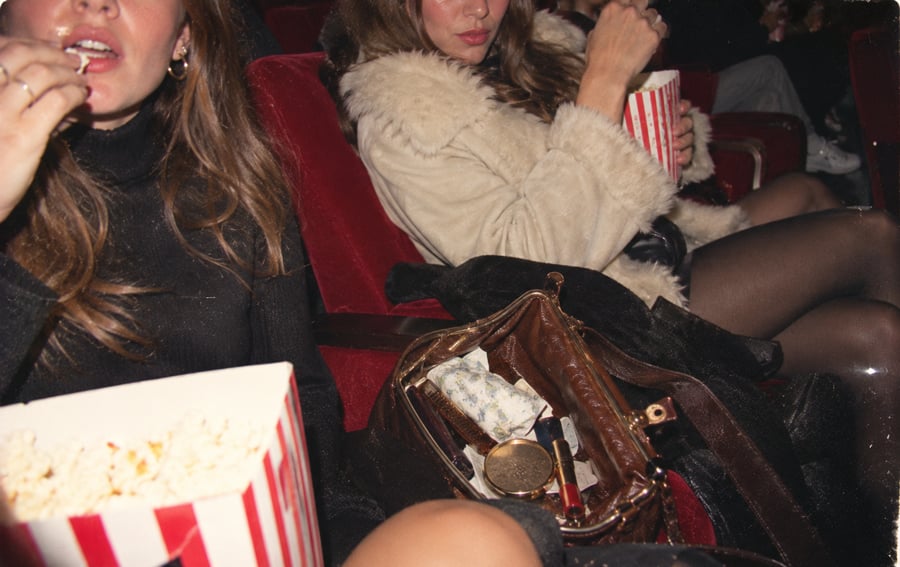 Two women sit in red theater seats, eating popcorn. One wears a fur coat and has a brown purse open on her lap, showing tissues and makeup items. The scene suggests they are watching a movie.